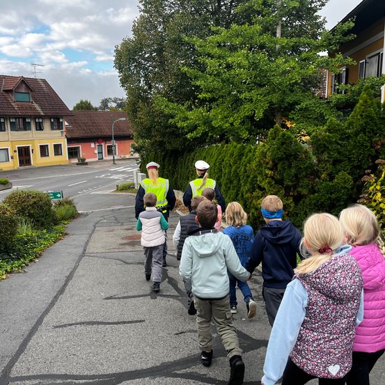 Eine Gruppe von Kindern läuft auf der Straße, begleitet von zwei Polizeibeamten. Die Kinder halten Händchen, und die Beamten tragen reflektierende Westen. Bäume und Gebäude befinden sich auf beiden Seiten der Straße.