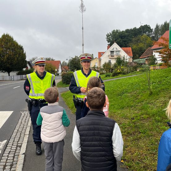 Zwei Polizeibeamte in neongelben Westen stehen auf der Straße und erklären einer Gruppe von Kindern Verkehrssicherheit. Die Kinder hören aufmerksam zu. Im Hintergrund sind Häuser und Bäume zu sehen.