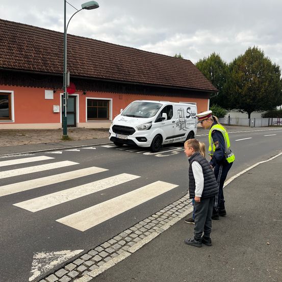 Ein Verkehrshelfer in einem Warnweste gibt zwei Kindern auf einem Zebrastreifen Anweisungen. Hinter ihnen steht ein weißer Lieferwagen mit einer Werbung für "Schaffer" an der Kreuzung.