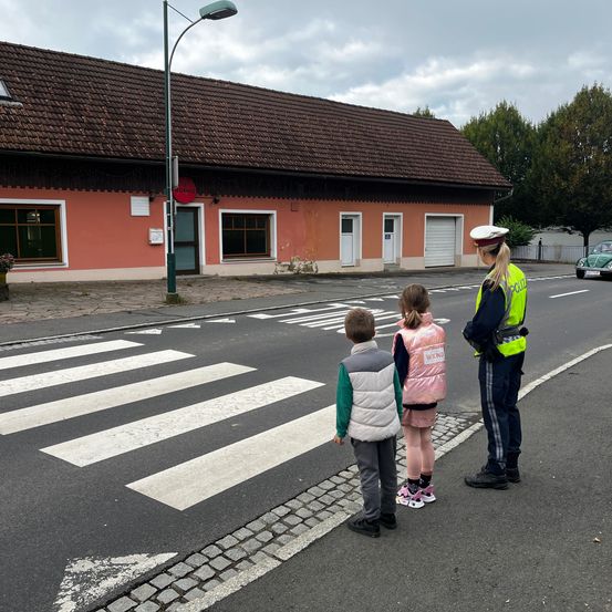 Ein Verkehrspolizist weist zwei Kinder an einem Zebrastreifen vor einem Gebäude mit mehreren Fenstern an.