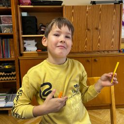 Ein junger Junge in einem gelben Sweatshirt hält eine orange Karotte und lächelt, sitzt in einem Klassenzimmer.