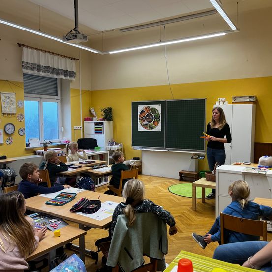 Eine Lehrerin steht vor einer Tafel im Klassenzimmer und hält einen orangefarbenen Bleistift. Mehrere Schüler sitzen an Tischen, einige von ihnen tragen Brillen. Der Raum hat Holzböden und gelbe Wände.