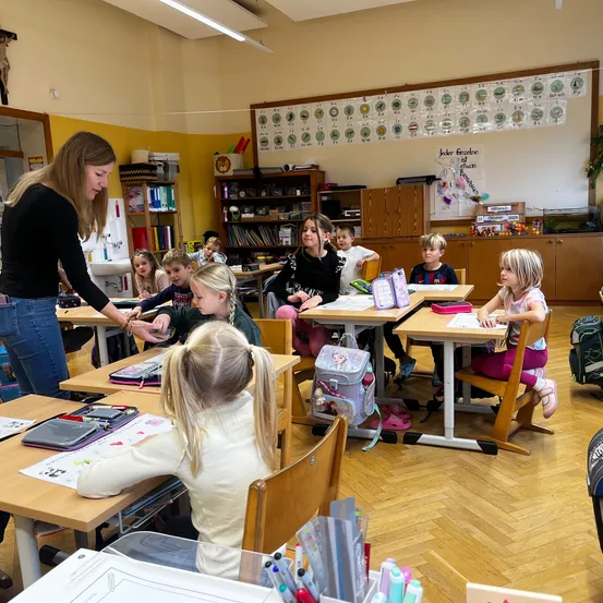 Eine Lehrerin hilft einer Schülerin in einem Klassenzimmer mit Schreibtischen, Rucksäcken und einer farbenfrohen Wand. Die Lehrerin trägt ein schwarzes Shirt und Jeans, und die Schüler sind mit ihren Aufgaben beschäftigt.