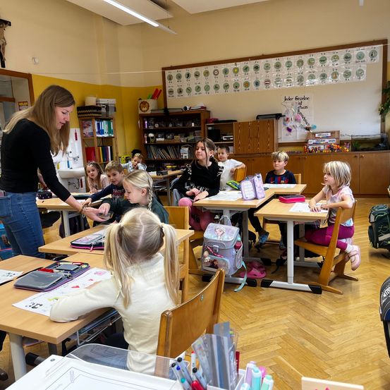 Eine Lehrerin hilft einer Schülerin in einem Klassenzimmer mit Schreibtischen, Rucksäcken und einer farbenfrohen Wand. Die Lehrerin trägt ein schwarzes Shirt und Jeans, und die Schüler sind mit ihren Aufgaben beschäftigt.