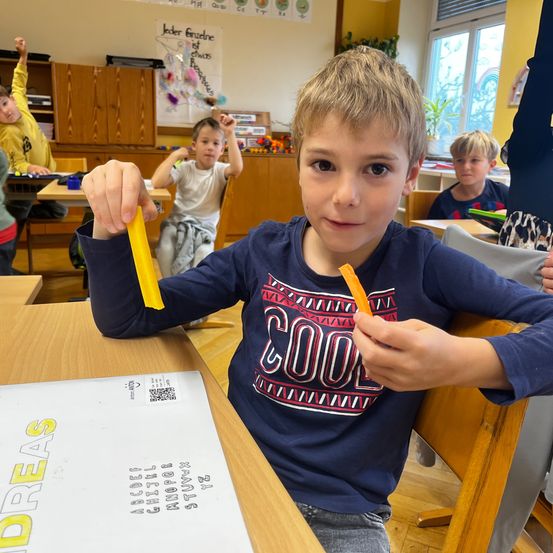 Ein junger Junge im Klassenzimmer hält zwei Papierstreifen in der Hand. Ein Blatt Papier mit dem Wort Ideen steht auf dem Tisch vor ihm.