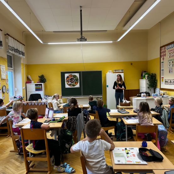 Ein Klassenzimmer mit gelben Wänden, Schüler sitzen an Tischen, eine Lehrerin steht neben einer Tafel, Projektor hängt an der Decke.