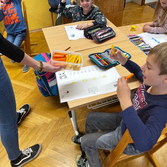Ein Klassenzimmer mit Kindern an Tischen, ein Junge isst Karotten, ein anderer hält einen Marker. Eine Lehrerin verteilt Karotten. Eine Mädchen lächelt im Hintergrund.