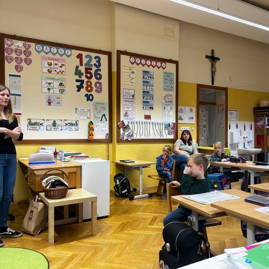 Ein Klassenzimmer mit Holzboden und gelben Wänden. Eine Lehrerin steht neben einer Tafel mit Zahlen und einem Kreuz an der Wand. Schüler sitzen an Tischen, einige tragen Jacken. Ein Kühlschrank und ein kleiner Tisch mit einem Korb sind im Raum.