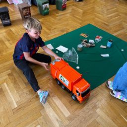 Ein Junge spielt mit einem Spielzeug-Müllwagen auf einem Holzboden. Verschiedene Müllgegenstände sind um ihn herum verstreut.