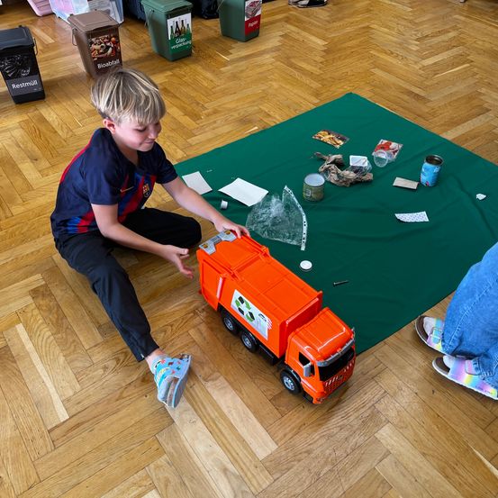 Ein Junge spielt mit einem Spielzeug-Müllwagen auf einem Holzboden. Verschiedene Müllgegenstände sind um ihn herum verstreut.
