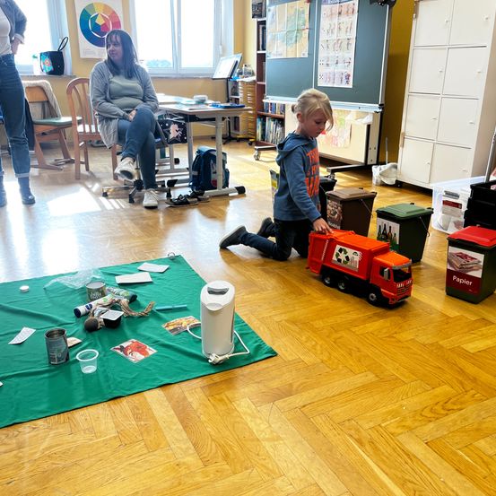 Ein junges Kind kniet neben einem Spielzeuglastwagen auf einem grünen Teppich. Eine Frau sitzt im Hintergrund. Auf dem Teppich liegen verschiedene Gegenstände wie ein Becher, eine Dose und eine Flasche.