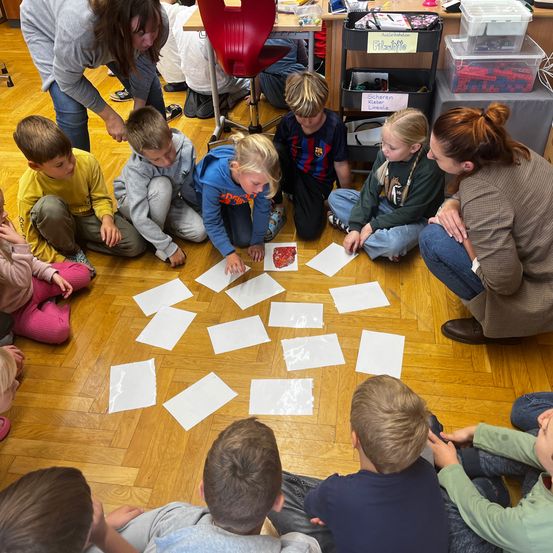 Kinder sitzen im Kreis auf dem Boden, mit Papier auf dem Boden und zwei Erwachsenen, die stehen. Eine Erwachsene hilft einem Kind mit einem Papier.
