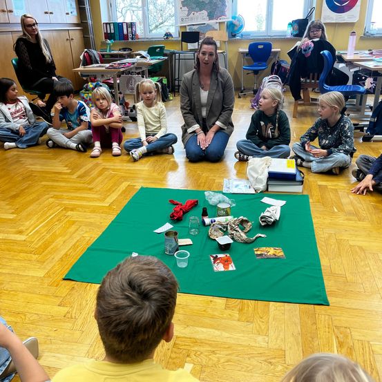 Eine Gruppe von Kindern und einer Frau sitzen auf einem Holzboden. Sie sind von verschiedenen Gegenständen auf einem grünen Tuch umgeben. Die Frau scheint die Kinder anzusprechen.