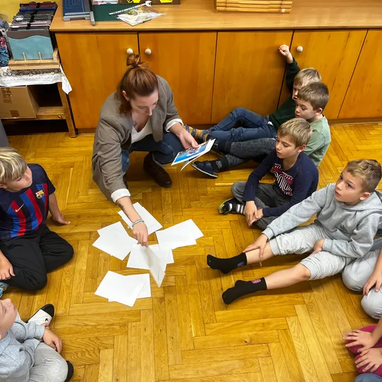 Eine Frau liest einem Jungenkreis auf dem Boden vor. Papiere sind auf dem Boden verstreut.
