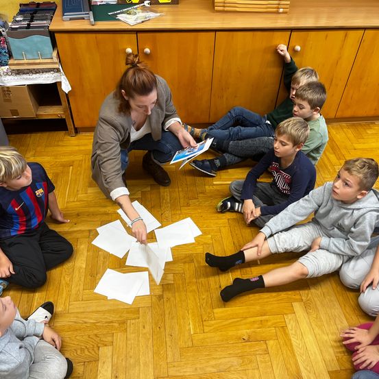 Eine Frau liest einem Jungenkreis auf dem Boden vor. Papiere sind auf dem Boden verstreut.