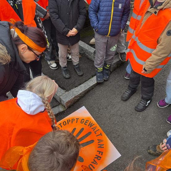 Menschen in orange Westen sind um ein Schild herum versammelt. Eine Frau vorne hat eine orange Kopfband. Das Schild sagt 'Stopp Gewalt An Frauen'.