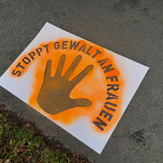 Ein orange-weißes Plakat auf dem Boden mit einem Handabdruck und dem Text 'STOPP GEWALT AN FRAUEN' in deutscher Sprache.