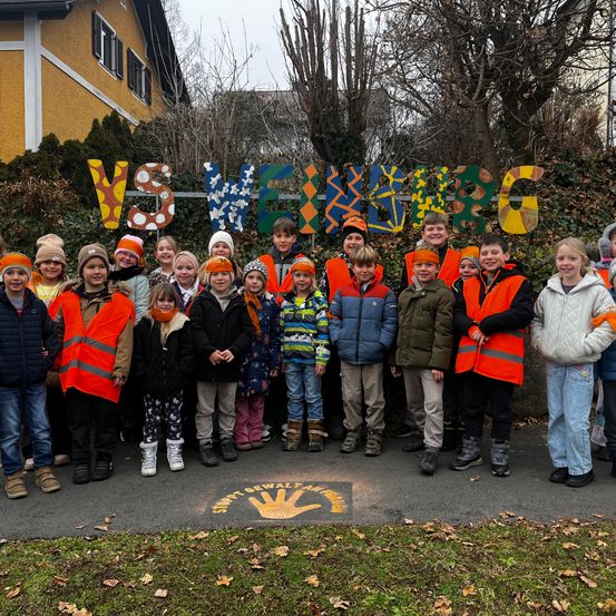 Eine Gruppe von Kindern in Warnwesten posiert für ein Foto vor einem Schild. Das Schild hat das Wort 'Weinberg' darauf. Die Kinder stehen auf einem Betonweg.
