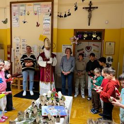 Eine Gruppe von Kindern und Erwachsenen versammelt sich um einen Mann, der als St. Nikolaus verkleidet ist. Sie stehen in einem Klassenzimmer mit einem Kreuz und Dekorationen an der Wand. Die Kinder halten Geschenke und scheinen zu singen.