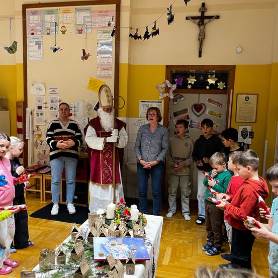 Eine Gruppe von Kindern und Erwachsenen versammelt sich um einen Mann, der als St. Nikolaus verkleidet ist. Sie stehen in einem Klassenzimmer mit einem Kreuz und Dekorationen an der Wand. Die Kinder halten Geschenke und scheinen zu singen.