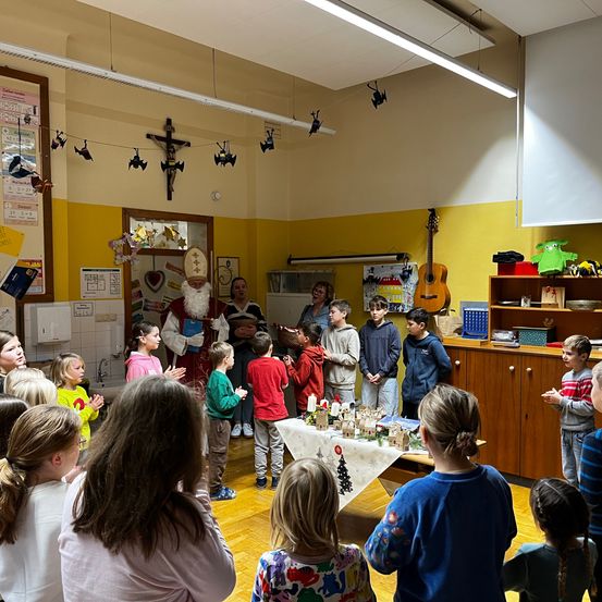 Eine Gruppe von Kindern versammelt sich um den Weihnachtsmann in einem Klassenzimmer, mit einem geschmückten Tisch und einer Lehrerin in der Nähe. Ein Kreuz und Halloween-Dekorationen hängen von der Decke.