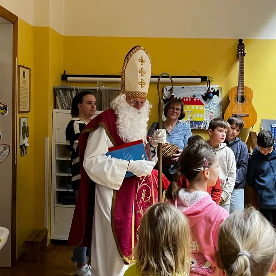 Ein Mann, der als St. Nikolaus verkleidet ist, steht in einem Raum mit Kindern. Er hält ein blaues Buch und einen Stab. Der Raum hat gelbe Wände und ein Regal mit Gegenständen. Zwei Gitarren sind an der Wand montiert.
