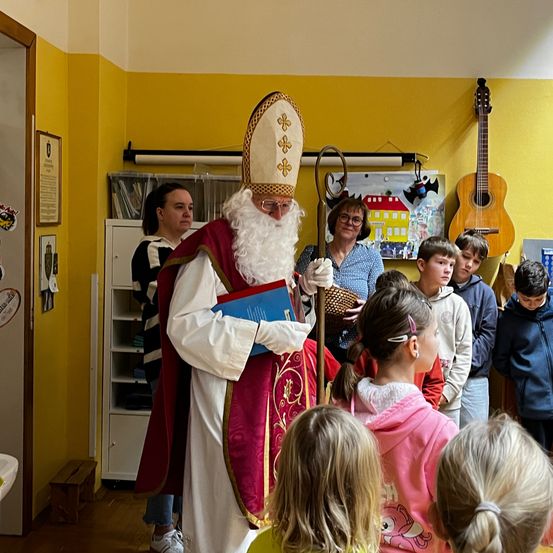 Ein Mann, der als St. Nikolaus verkleidet ist, steht in einem Raum mit Kindern. Er hält ein blaues Buch und einen Stab. Der Raum hat gelbe Wände und ein Regal mit Gegenständen. Zwei Gitarren sind an der Wand montiert.