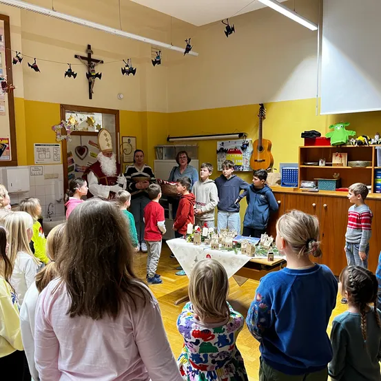 Eine Gruppe von Kindern versammelt sich um einen Tisch im Klassenzimmer und beobachtet ein Modelldorf. Eine Frau in einem blauen Shirt und Brille steht in der Nähe. Oben schmücken ein Kreuz und Halloween-Dekorationen die gelbe Wand.