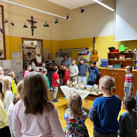 Eine Gruppe von Kindern versammelt sich um einen Tisch im Klassenzimmer und beobachtet ein Modelldorf. Eine Frau in einem blauen Shirt und Brille steht in der Nähe. Oben schmücken ein Kreuz und Halloween-Dekorationen die gelbe Wand.