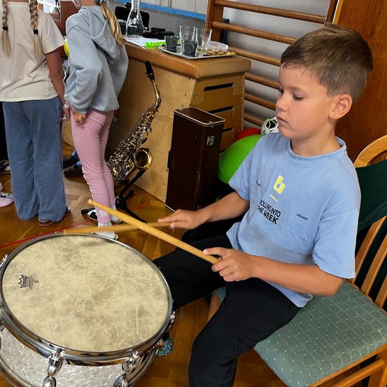 Ein kleiner Junge spielt mit zwei Schlägeln Schlagzeug in einem Klassenzimmer. Hinter ihm beobachtet ein Mädchen. Im Hintergrund sind verschiedene Musikinstrumente und Turngeräte zu sehen.