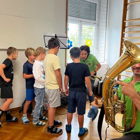 Eine Gruppe von Jungen steht um einen Notenständer und eine Frau mit einer Tuba herum. Sie sind in einem Raum mit Holzböden und weißen Wänden.