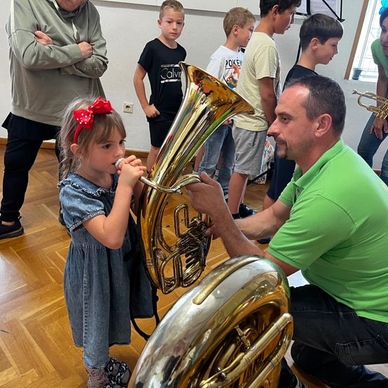 Ein kleines Mädchen bläst in eine Tuba, während ein Mann in Grün das Instrument hält. Andere Kinder schauen zu.