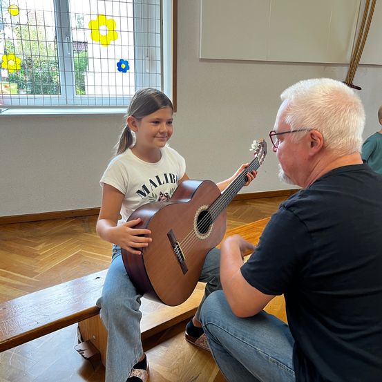 Ein junges Mädchen mit einer Gitarre sitzt auf einer Bank, lächelt, während ein älterer Mann mit einer Brille ihr hilft. Hinter ihnen ist ein Fenster mit Blumendekorationen.