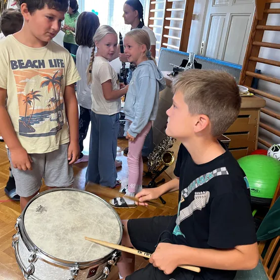 Mehrere Kinder befinden sich in einem Musikzimmer, wobei ein Junge ein Schlagzeug spielt und ein anderer Junge in einem gelben T-Shirt zuschaut. Ein Mädchen schaut zu, während ein anderes Mädchen eine Saxophon hält.