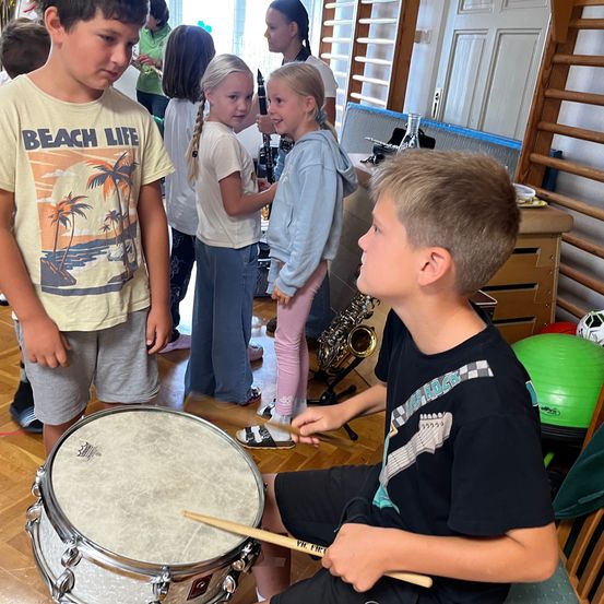 Mehrere Kinder befinden sich in einem Musikzimmer, wobei ein Junge ein Schlagzeug spielt und ein anderer Junge in einem gelben T-Shirt zuschaut. Ein Mädchen schaut zu, während ein anderes Mädchen eine Saxophon hält.