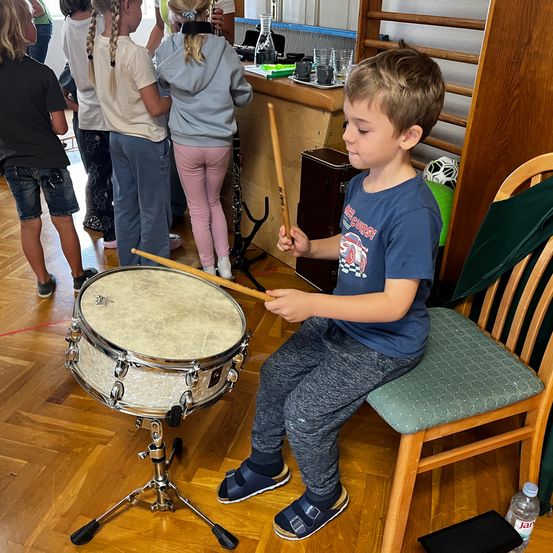 Ein kleiner Junge sitzt auf einem Stuhl und spielt die Trommeln in einem Musiksaal. Andere Kinder beobachten.
