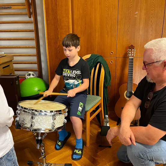 Ein junger Junge spielt eine Trommel und ein älterer Mann beobachtet ihn. Sie sind in einem Raum mit Holzwänden und einer Gitarre in der Nähe.