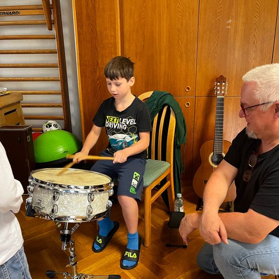 Ein junger Junge spielt eine Trommel und ein älterer Mann beobachtet ihn. Sie sind in einem Raum mit Holzwänden und einer Gitarre in der Nähe.