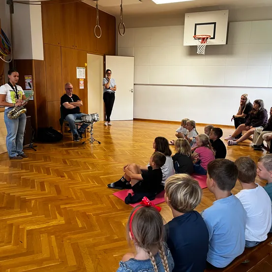 Eine Gruppe von Kindern sitzt auf dem Boden und beobachtet eine Frau, die Saxophon spielt, und einen Mann, der Schlagzeug spielt. Der Raum hat Holzböden und einen Basketballkorb an der Wand.