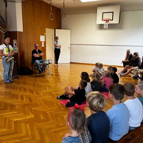 Eine Gruppe von Kindern sitzt auf dem Boden und beobachtet eine Frau, die Saxophon spielt, und einen Mann, der Schlagzeug spielt. Der Raum hat Holzböden und einen Basketballkorb an der Wand.