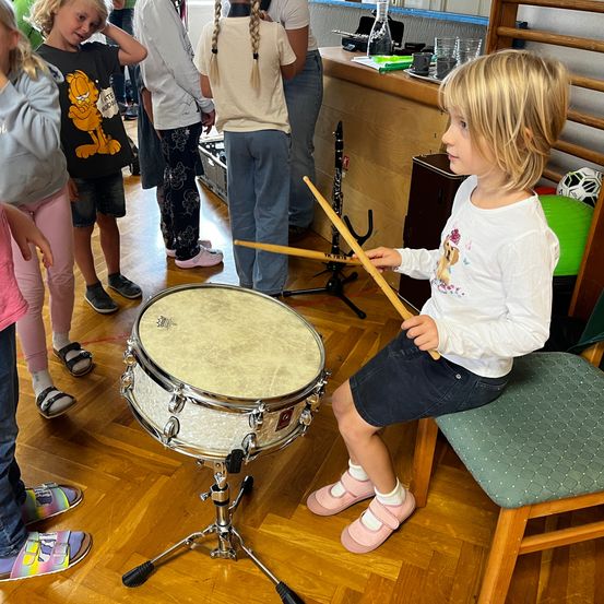 Ein kleines Mädchen sitzt und spielt ein Schlagzeug mit Schlägeln, umgeben von Kindern, die zusehen. Der Raum ist gut beleuchtet, und der Boden ist aus Holz.