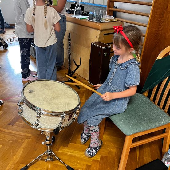 Ein kleines Mädchen mit Zöpfen und einer roten Schleife spielt ein Schlagzeug, sitzt auf einem Stuhl in einem Musikraum. Andere Kinder beobachten im Hintergrund.