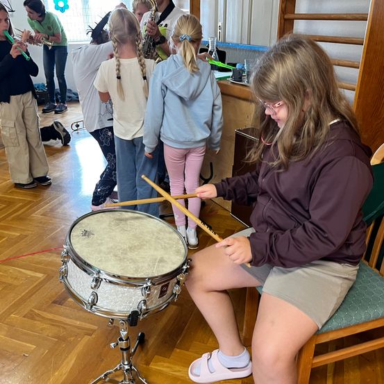 Ein junges Mädchen spielt die Trommel, während andere verschiedene Instrumente in einem Raum spielen. Sie sitzt auf einem Stuhl und hält Trommelstöcke. Hinter ihr stehen einige Leute und halten Musikinstrumente.