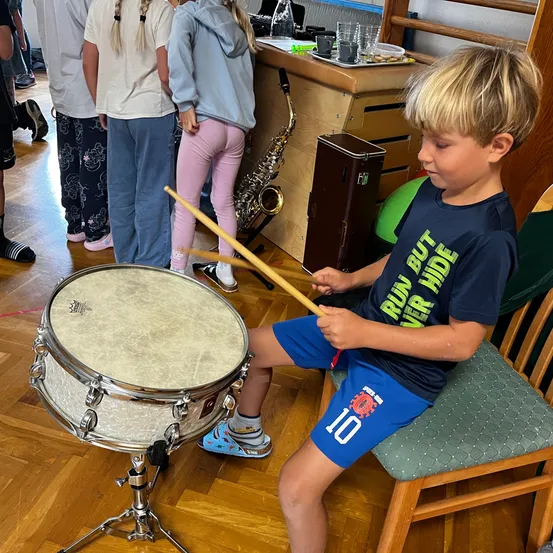 Ein junger Junge spielt die Trommel, während er auf einem Stuhl sitzt. Andere beobachten von hinten. Der Raum hat einen Holzboden und eine Holztheke mit einer Saxophon und Tassen.