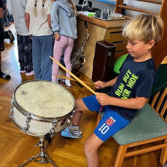 Ein junger Junge spielt die Trommel, während er auf einem Stuhl sitzt. Andere beobachten von hinten. Der Raum hat einen Holzboden und eine Holztheke mit einer Saxophon und Tassen.