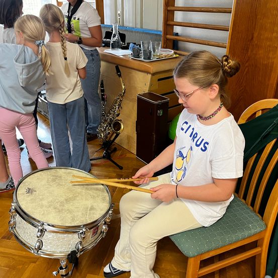 Ein Mädchen spielt mit Schlägeln eine Trommel in einem Musikraum, mit einer Klarinette und einer Lehrerin in der Nähe. Ein anderes Mädchen beobachtet.