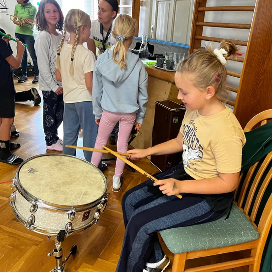Eine Gruppe junger Mädchen spielt in einem Raum Instrumente. Ein Mädchen vorne spielt die Trommel, während andere zuschauen.