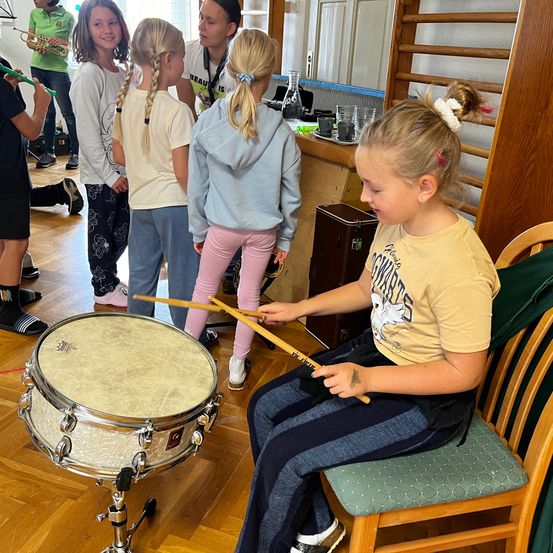 Eine Gruppe junger Mädchen spielt in einem Raum Instrumente. Ein Mädchen vorne spielt die Trommel, während andere zuschauen.