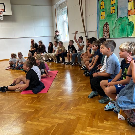 Eine Gruppe von Kindern sitzt kreisförmig in einem Raum mit Holzböden. Einige sitzen auf Yoga-Matten. Eine Frau steht davor. Ein Basketballkorb und ein farbenfrohes Wandbild sind im Hintergrund.