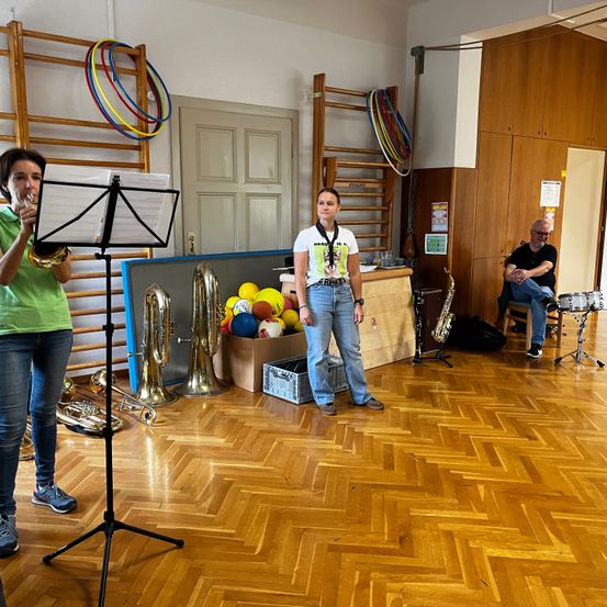 Eine Frau steht und spielt Trompete, während eine andere Frau sie ansieht. Musikinstrumente sind im Raum verteilt, und ein Mann sitzt auf einem Hocker vor einem Schlagzeug.